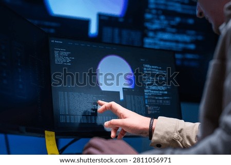 A serious young security operations analyst points at the terminal explaining the output inside a modern security operations centre after hours. The scene shows hunting for digital threats.