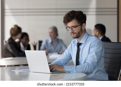 Serious Young Startup Leader Guy Working On Project In Co-working Space. Millennial Businessman In Glasses Using Laptop Computer In Office With Corporate Team In Background