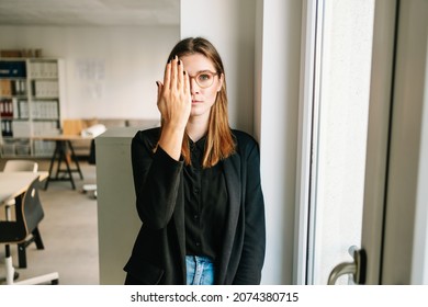Serious Young Woman With A Deadpan Expression Covering One Eye With Her Hand As She Stares At The Camera Indoors In A Spacious Office