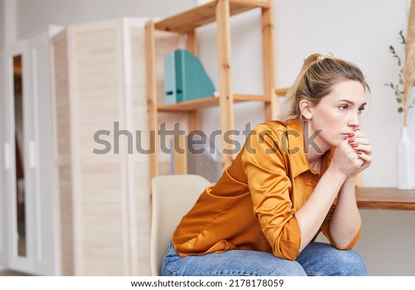 Serious young\
woman in despair sitting on chair in living room and leaning head\
on hand while thinking of own\
life