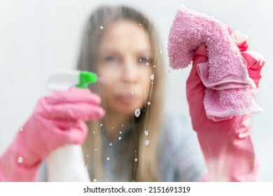 A serious young woman in pink protective gloves looks attentively while washing a dirty window with spray foam and a rag. Females hands hold a rag and wipes the transparent glass. Cleaning windows.