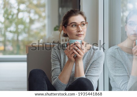 Serious young woman sitting daydreaming with mug of coffee as she stares out of a large window with faraway thoughtful expression, reflection on the glass and copyspace