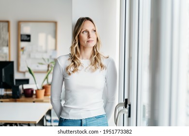 Serious young woman standing deep in thought looking through a window in a high key office with copyspace looking up with a contemplative expression