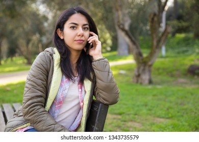 Serious young woman talking on mobile phone in park and looking at camera. Sad attractive girl attentively listening to friend on phone. Communication concept