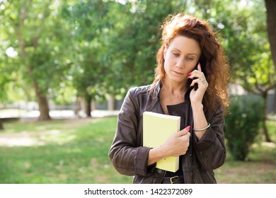 Serious Young Woman Talking On Phone. Pensive Pretty Brunette Walking On Campus With Book And Making Phone Call. Communication Concept