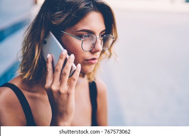 Serious young woman in trendy eyewear concentrated on information getting during phone talk with customer support operator, thoughtful hipster girl calling to friend standing near publicity area