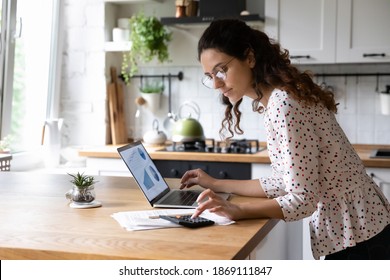 Serious young woman wearing glasses calculating finances, household expenses, confident businesswoman working with project statistics, using laptop and calculator, standing in kitchen at home - Shutterstock ID 1869111847