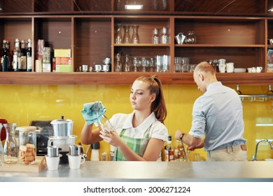 Serious Young Woman Working In Coffeeshop Or Restaurant, She Is Wiping Glass Jars When Getting Ready For Opening In The Morning