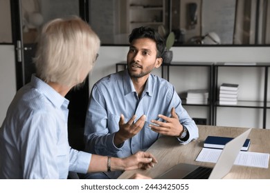 Serious younger Indian professional man talking to elder colleague woman at workplace, discussing project, collaboration, management strategy. Business coworkers speaking at office table - Powered by Shutterstock