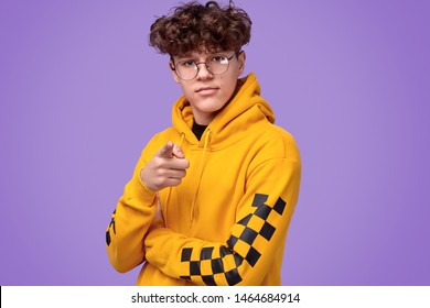 Serious youngster with curly hair wearing nerdy glasses and yellow hoodie and pointing at camera while standing against violet background - Powered by Shutterstock