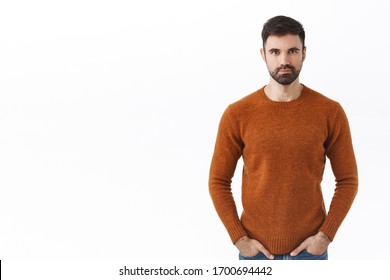 Serious-looking assertive bearded man, family guy in sweatshirt, hold hands in pockets and look camera determined with pleased, confident smile, standing white background on right side