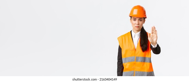 Serious-looking disappointed asian female architect, construction manager at working area wearing safety helmet, showing stop gesture, prohibit action, forbid trespassing, white background - Shutterstock ID 2163404149