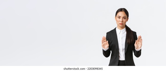 Serious-looking displeased asian female manager, businesswoman in black suit stop action, prohibit or restrict something, raising hands in forbid gesture, saying no or enough, white background - Powered by Shutterstock