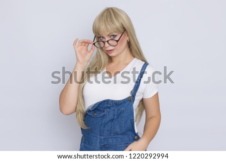 Seriously blond woman in jeans overalls holding glasses on her face over grey background