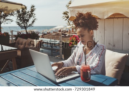 Serous busy young biracial lady in traditional African chemise is sitting with laptop in street cafe nearshore with jar of reddish cold lemonade and working on her modern laptop, sunny summer day
