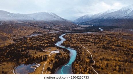 A serpentine turquoise river winds through a valley with rich autumnal foliage, flanked by towering, snow-capped mountains under a cloudy sky. - Powered by Shutterstock