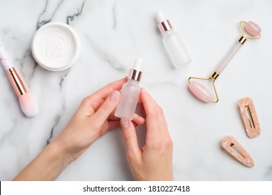 Serum Bottle Dropper In Female Hands Over Beauty Products For Face Treatment On Marble Table. Flat Lay, Top View. Face Or Body Skin Care Concept.