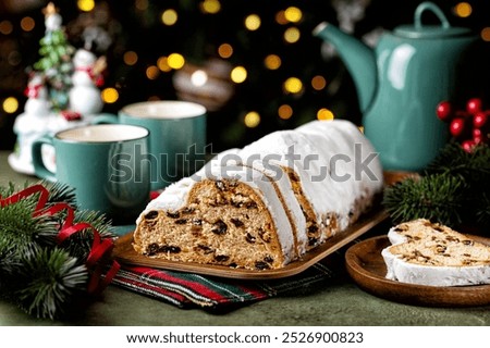 Served festive table with Stollen cake, tea cups and pot. German Christmas dessert, bread chock full of dried fruit, candied peel, and almonds. Festive lights, tree brunch and decoration.