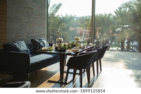 served table with sofa and chairs for a large company against the background of a large window overlooking the palm trees