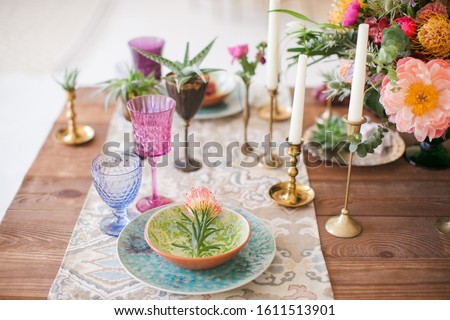 Served table for a wedding dinner in boho chic style. Decor from fresh flowers of peonies, roses, orchids and Leucospermum conocarpodendron. Crockery plates, glasses and candles. Wooden table.