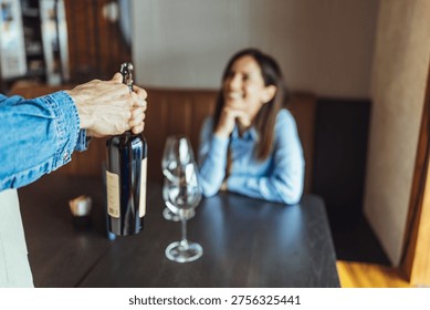 Server opens a wine bottle while a smiling woman watches at a cozy restaurant table, two wine glasses ready for a relaxed, romantic date and friendly hospitality in a warm interior setting.