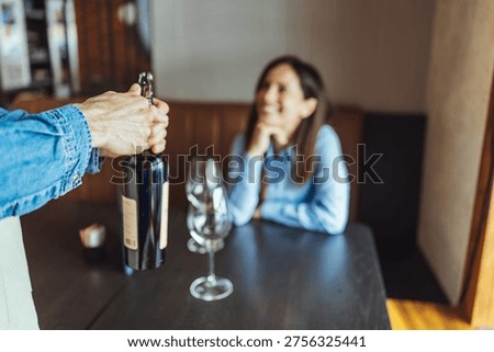 Server opens a wine bottle while a smiling woman watches at a cozy restaurant table, two wine glasses ready for a relaxed, romantic date and friendly hospitality in a warm interior setting.