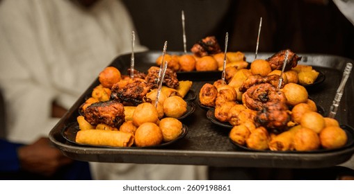 Server presenting assorted Nigerian small chops platter, featuring puff-puff, grilled chicken, samosas, and spring rolls at a social event in Lagos