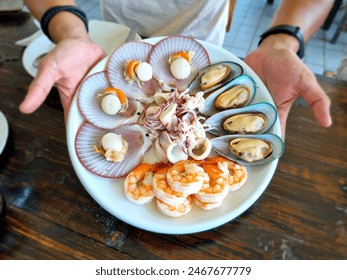 A server presents a platter of fresh seafood at a restaurant on Koh Kood island, Trat Province. The dish features scallops, mussels, squid, and prawns, highlighting the local catch.