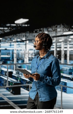 Server room admin using tablet and headset microphone to provide tech support on industrial platform in server farm. Data center IT expert resolves backend issues, coordinating remote teams.