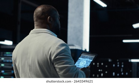 Server room software developer troubleshooting errors using software on laptop. African american IT professional walking in data center fixing bugs using notebook, camera B