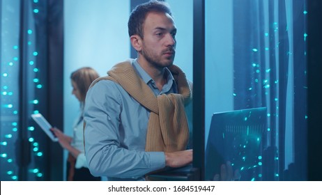 Server tech support with headset performing data operations in server room. His female co-worker administrator checking rack server cabinets updating hardware with digital tablet. Teamwork.