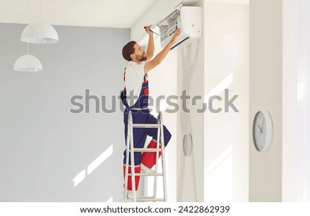 Service worker stands on ladder in modern flat or office, sets up new AC, repairs some parts, or removes dirty purifier. Air conditioning installation, full warranty repair and maintenance concept