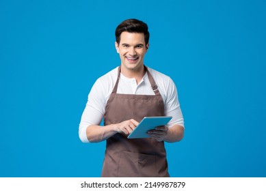 Serviced minded Caucasian male waiter with apron holding tablet in his hands while standing on blue background in light studio