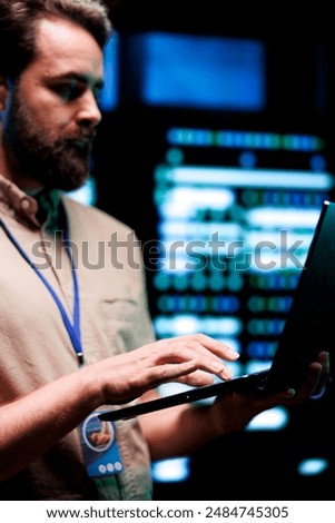 Serviceman doing system analysis, typing data on laptop of needed adjustments in supercomputers server farm housing advanced data storage infrastructure supporting critical IT workloads, close up