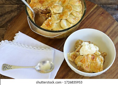 Serving Of Apple Pie With Dollop Of Cream In A Bowl Next The Pie In A Glass Bowl.  A Serviette And Spoon Sits Next To The Bowl On A Wooden Board.