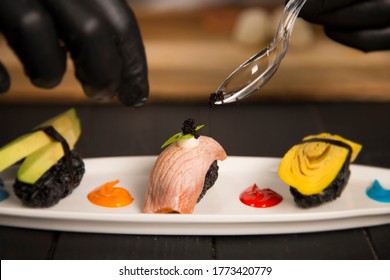 Serving Black Tobiko Caviar On Japanese Nigiri Sushi Sake With Salmon Tataki On White Plate. Chef Hands In Protection Gloves Using Tea Spoon. Pan Asian Restaurant Food Decoration Process.