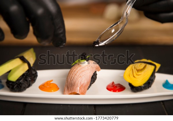 Serving black Tobiko caviar on Japanese Nigiri\
Sushi Sake with salmon Tataki on white plate. Chef hands in\
protection gloves using tea spoon. Pan Asian Restaurant Food\
decoration process.