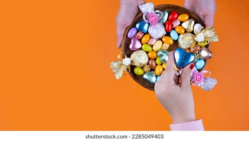 Serving candies, mother holding bowl and serving candies. Top view isolated orange background, copy space. Ramadan or Ramazan feast celebration concept idea. Girl hand taking one wrapped chocolate.