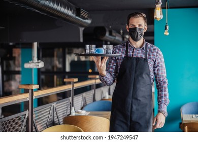 Serving Coffee And Water During The Corona Virus. Portrait Of A Male Waiter In A Plaid Shirt And Apron With A Protective Face Mask Walking Around A Cafe Carrying A Tray Of Coffee And Water