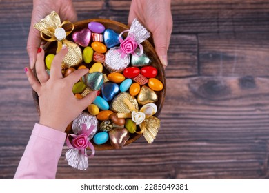 Serving confectionery, top view image of woman hand holding serving confectionery. Child girl taking one of the sweets. Wrapped chocolates, almond dragee. Wooden background, copy space.