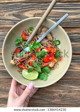 Serving food on rustic wooden table. Soba noodles with colorful vegetables and chicken strips. Top view. Woman’s hand holding the plate.