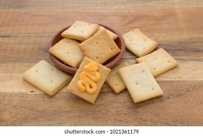 A Serving Of Hard Bread Crackers In A Red Clay Bowl With One In Front Covered With Canned Cheese On A Wood Table.