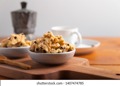 A Serving Of Homemade Cookie Dough On A Wooden Table, With A Wooden Spoon. With Coffee In The Background.