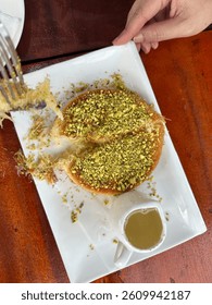 A serving of Kunafa, a Middle Eastern dessert topped with crushed pistachios, served with a side of sweet syrup for pouring.