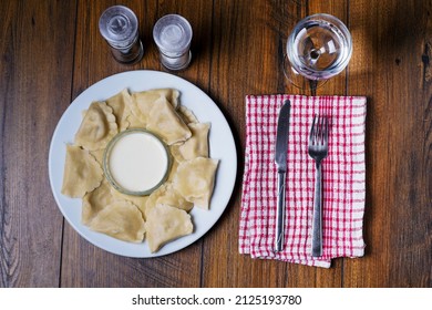 Serving Of Polish Dumplings With Sour Cream On White Plate And Glass Of Water And Salt And Black Pepper Shaker. Simple Meal On A Wooden Table, Top Down View.