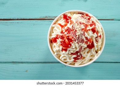 Serving of speciality German vanilla spaghetti ice-cream sundae in a takeaway tub topped with fresh strawberry coulis or sauce over blue wood with copyspace for menu advertising