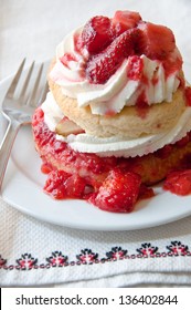 A serving of strawberry shortcake sits on a plate. Under the plate is a white napkin with fancy red and black embroidery.