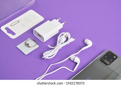 A Set Of Accessories For A Smartphone, Phone, Charger, Headphones, A Ring-holder, Adapters For SIM Cards On A Purple Background Copy Space Top View