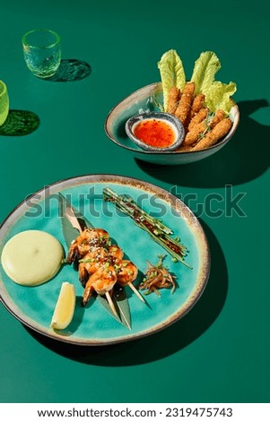 Set of Asian appetizers: cheese tempura with sweet chili sauce and shrimp skewers with wasabi sauce. Presented on a solid green background with harsh shadows from glassware.