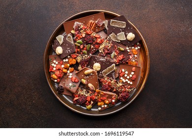 Set of assorted broken handmade chocolate bars pieces with nuts, dried berries and marmalade in a plate over dark brown background. Overhead view, copy space